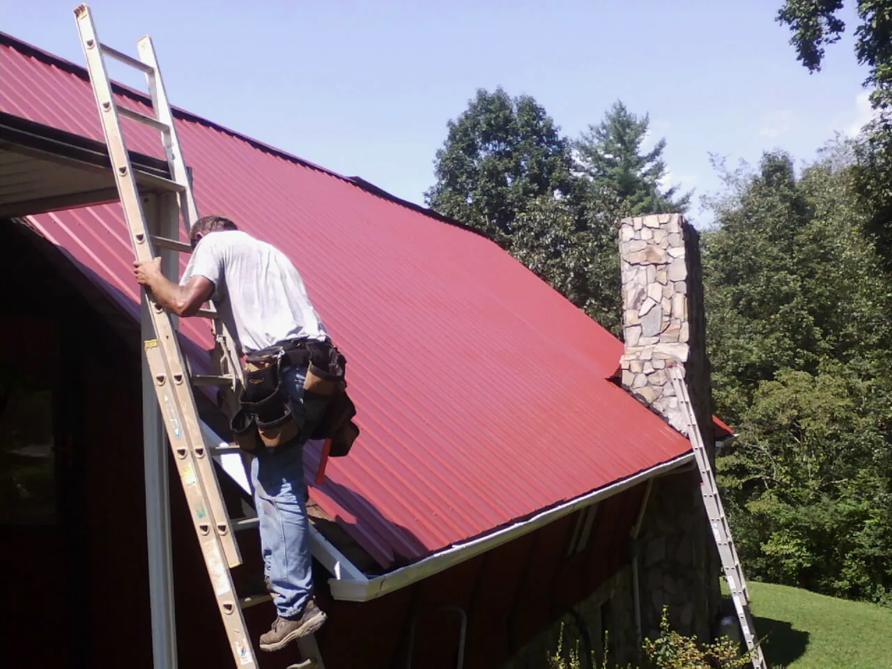 Roofer on a ladder beside a red metal roof and stone chimney — Soffit & Fascia Repair by Roofing Brothers in Olanta